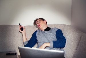 Young man asleep on sofa with remote control and laptop